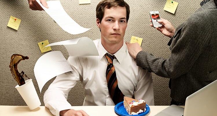 stern-faced man sitting in a cubicle looking at camera, two people offscreen reach their hand into view to hand him papers